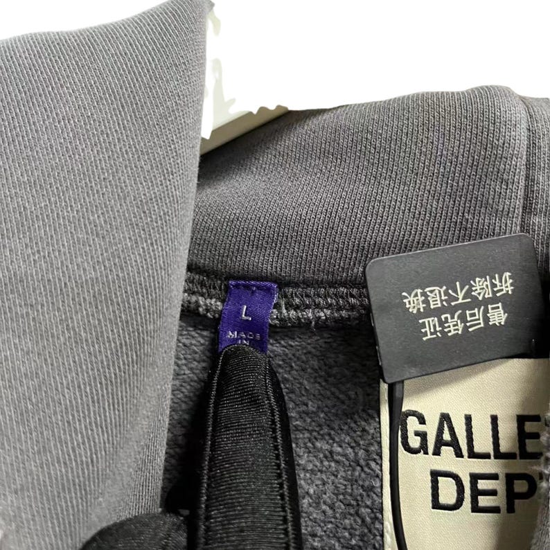 May include: Close-up of a gray hooded sweatshirt with a tag that reads "GALLE DEP". The tag also has Chinese characters and a size "L" label. The sweatshirt is made of a soft, textured fabric.