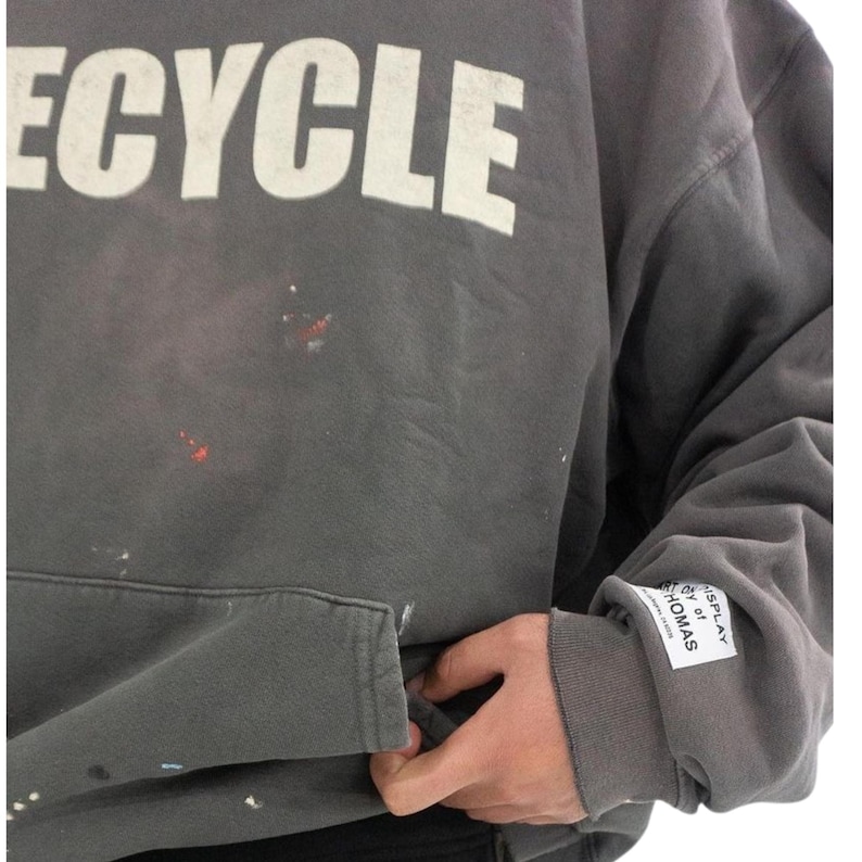 May include: Gray hoodie with the word "RECYCLE" in large white letters across the chest. The hoodie has a front pocket and a label on the sleeve that reads "DISPLAY BY THOMAS". There are paint splatters on the front.
