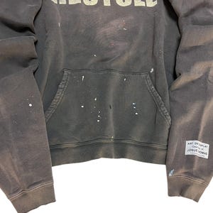May include: Dark gray hoodie with white paint splatters and the word "RECYCLED" across the chest. Features a front pocket and a small tag on the sleeve that reads "ART ON DISPLAY Country of JOSUE THOMAS."
