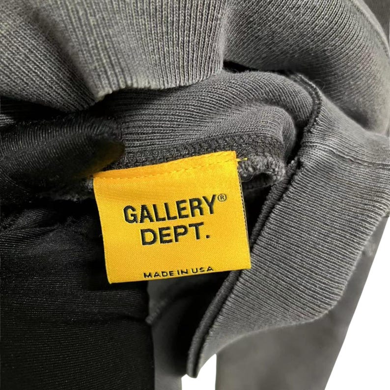 May include: Close-up of a gray sweatshirt with a yellow tag. The tag reads "GALLERY DEPT." in black capital letters, with "MADE IN USA" below. The sweatshirt is made of a ribbed fabric.