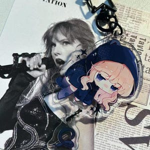 May include: A clear acrylic keychain featuring a cartoon character in a blue hooded outfit. The keychain is attached to a black star-shaped clasp. The background includes a black and white image of a singer and the word "REPUTATION".