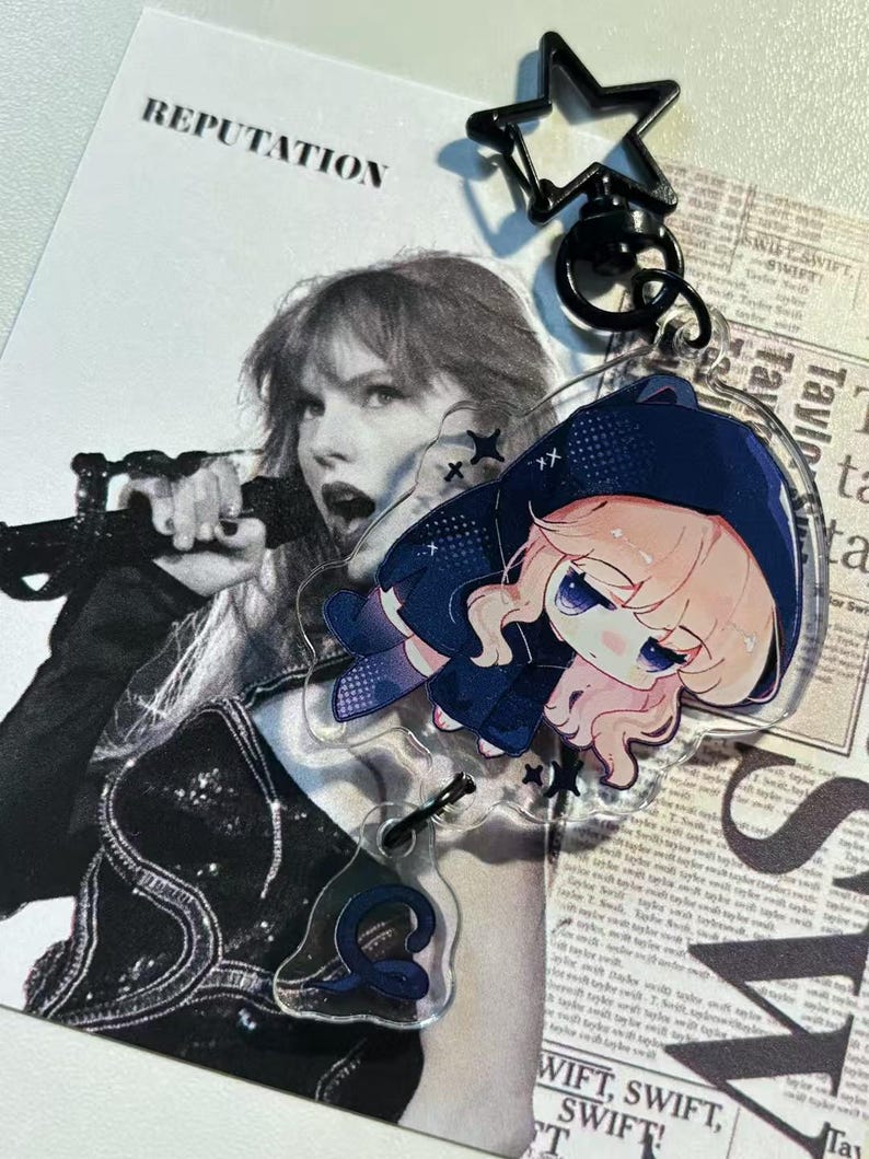 May include: A clear acrylic keychain featuring a cartoon character in a blue hooded outfit. The keychain is attached to a black star-shaped clasp. The background includes a black and white image of a singer and the word "REPUTATION".