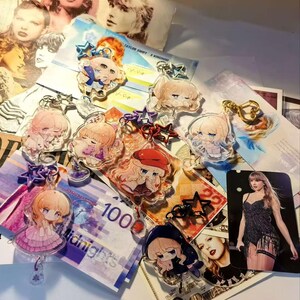 May include: A collection of acrylic keychains featuring anime-style illustrations of a blonde character in various outfits. The keychains are scattered on a surface with images of a singer and currency.