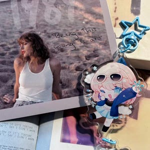 May include: A clear acrylic charm featuring an anime-style character with pink hair, sunglasses, and a blue outfit. The charm is attached to a blue star-shaped keychain. A photo with the text "Being this young is art" is in the background.