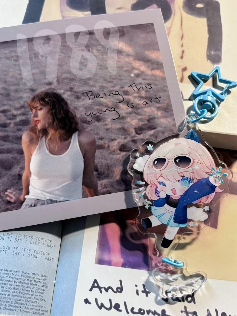 May include: A clear acrylic charm featuring an anime-style character with pink hair, sunglasses, and a blue outfit. The charm is attached to a blue star-shaped keychain. A photo with the text "Being this young is art" is in the background.