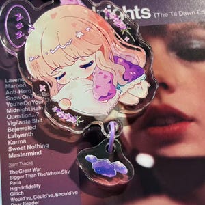 May include: A clear acrylic keychain featuring a sleeping anime-style character with light pink hair and purple accents. The keychain has a purple clasp and a smaller charm with a cloud design. The background is a purple album cover.
