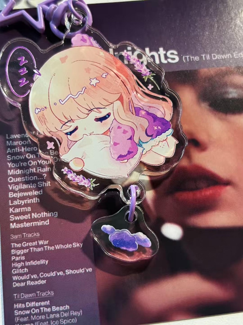May include: A clear acrylic keychain featuring a sleeping anime-style character with light pink hair and purple accents. The keychain has a purple clasp and a smaller charm with a cloud design. The background is a purple album cover.
