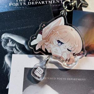 May include: A clear acrylic keychain featuring a cartoon character with light brown hair and purple eyes. The keychain has a star-shaped clasp and a small bottle charm. The background includes printed materials with the text "THE TORTURED POETS DEPARTMENT".
