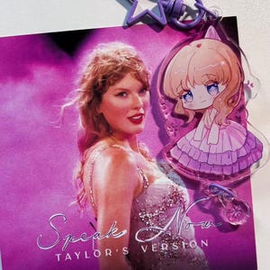 May include: A purple star-shaped keychain and a clear acrylic charm featuring an anime-style character are attached to a card with the text "Speak Now TAYLOR'S VERSION" and a photo of a woman in a sparkly dress. The background is a vibrant pink.