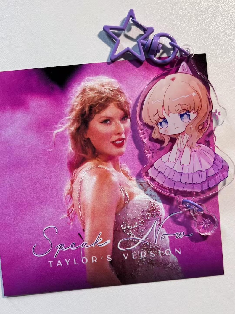 May include: A purple star-shaped keychain and a clear acrylic charm featuring an anime-style character are attached to a card with the text "Speak Now TAYLOR'S VERSION" and a photo of a woman in a sparkly dress. The background is a vibrant pink.
