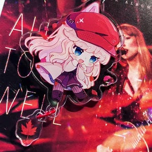 May include: A clear acrylic keychain featuring a cartoon character with light pink hair and a red hat. The keychain has a red maple leaf charm and is attached to a red metal ring. The background is a red album cover.