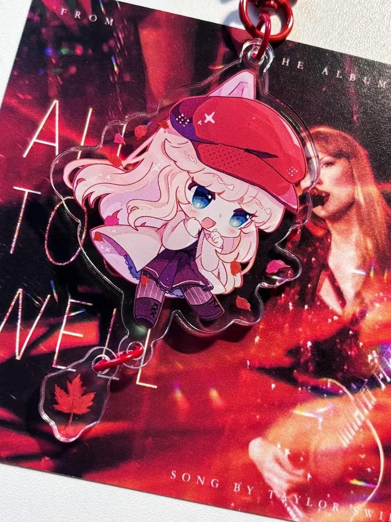 May include: A clear acrylic keychain featuring a cartoon character with light pink hair and a red hat. The keychain has a red maple leaf charm and is attached to a red metal ring. The background is a red album cover.