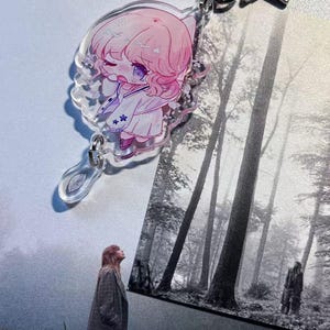 May include: A clear acrylic keychain featuring a pink-haired anime-style character with a white outfit and star accents. The keychain has a silver star charm and a leaf-shaped charm. The word "folklore" is printed at the top.