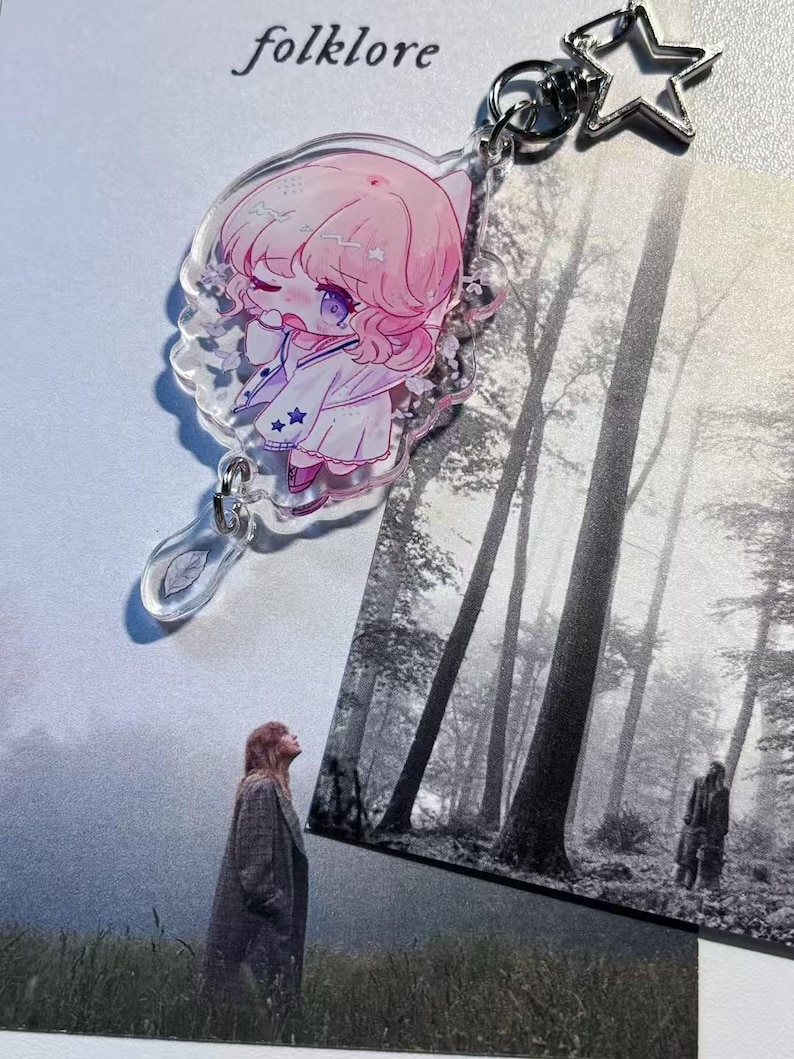May include: A clear acrylic keychain featuring a pink-haired anime-style character with a white outfit and star accents. The keychain has a silver star charm and a leaf-shaped charm. The word "folklore" is printed at the top.