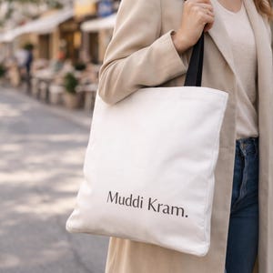 Könnte beinhalten: Eine weiße Canvas-Tragetasche mit schwarzen Trägern und dem Text "Muddi Kram." Die Tasche wird von einer Person getragen, die einen beigen Mantel und blaue Jeans trägt. Der Hintergrund zeigt eine verschwommene Straßenszene.