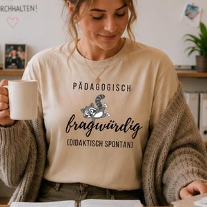 Może przedstawiać: Beżowa koszulka z niemieckim napisem "P&Auml;DAGOGISCH fragw&uuml;rdig (DIDAKTISCH SPONTAN)" i ilustracją szopa pracza z kresk&oacute;wki. Szop pracz jest szary i biały z paskowanym ogonem. Koszulka jest wykonana z miękkiego materiału.