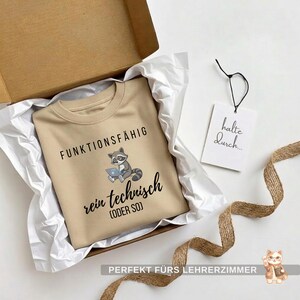 May include: A tan sweatshirt with the words "FUNKTIONSF&Auml;HIG rein technisch (ODER SO)" and a raccoon graphic. The sweatshirt is in a cardboard box with white tissue paper. A tag with "halte durch..." is attached.