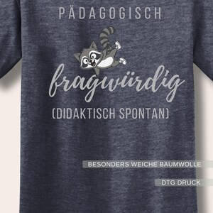 Może przedstawiać: Granatowa koszulka z niemieckimi słowami "P&Auml;DAGOGISCH fragw&uuml;rdig" i ilustracją szopa pracza z kresk&oacute;wki. Tekst "(DIDAKTISCH SPONTAN)" znajduje się pod gł&oacute;wnym tekstem. Koszulka zawiera r&oacute;wnież tekst "BESONDERS WEICHE BAUMWOLLE" i "DTG DRUCK".
