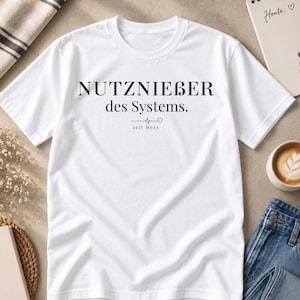 Könnte beinhalten: Ein weißes T-Shirt mit kurzen Ärmeln und dem schwarzen Schriftzug NUTZNIEßER des Systems. Unter dem Text befindet sich eine kleine Herzschlaglinie, ein Herzsymbol und das Wort seit Merz in einer kleineren Schriftgröße gedruckt.