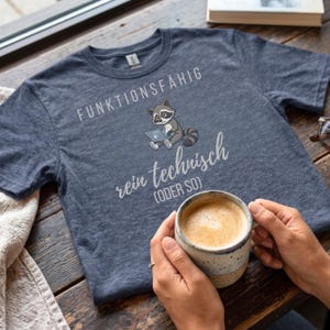 May include: A heather navy blue t-shirt with the words "FUNKTIONSF&Auml;HIG" and "rein technisch (ODER SO)" printed in white. A cartoon raccoon is depicted using a laptop. The shirt is on a wooden surface.