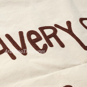 Puede incluir: Primer plano de una tela color crema con texto bordado en marr&oacute;n. El texto dice "EVERY" en letras grandes y estilizadas. El nombre de la tienda Etsy "ArtMinaTreasures" est&aacute; bordado debajo.