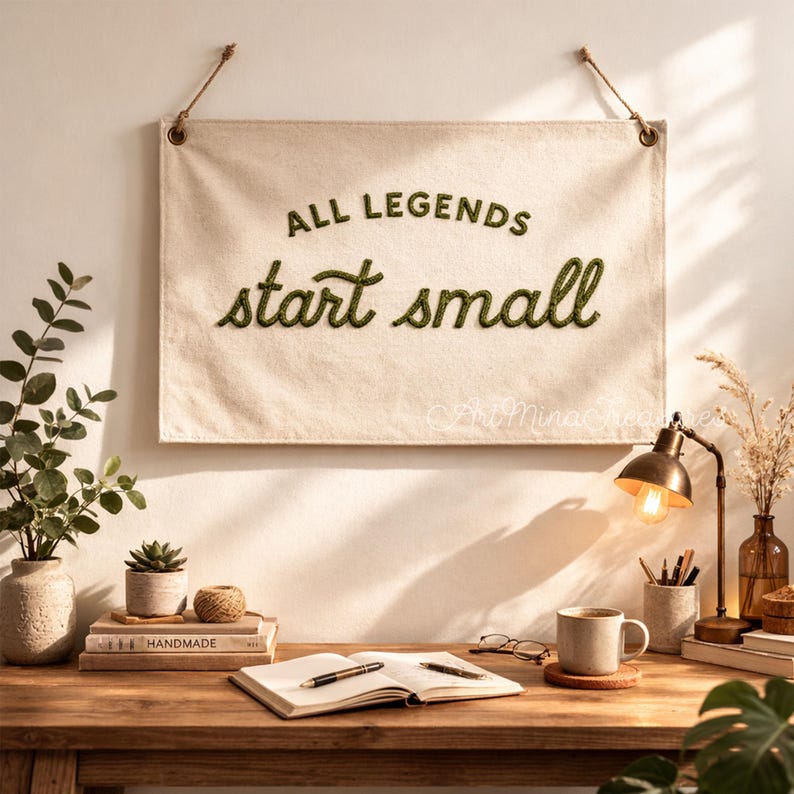 Puede incluir: Un tapiz de pared de lona color crema con el texto "ALL LEGENDS start small" en letras verdes. La pancarta est&aacute; suspendida por una cuerda y cuelga sobre un escritorio de madera con una l&aacute;mpara, una taza de caf&eacute; y un cuaderno abierto.