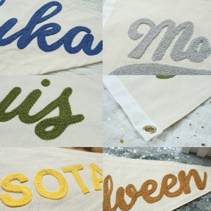 Puede incluir: Primer plano de varias pancartas de lona blanquecina con texto bordado en varios colores. Las pancartas presentan palabras como "Lika", "Mo", "Louis", "Sota" y "Halloween". Cada pancarta tiene un borde blanco y un ojal dorado.