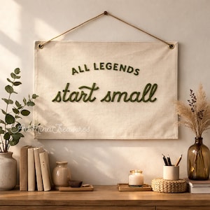 Puede incluir: Un tapiz de pared de lona beige con el texto "ALL LEGENDS start small" en verde. La pancarta est&aacute; suspendida por una cuerda marr&oacute;n. La imagen tambi&eacute;n muestra una mesa de madera con libros, jarrones y una vela.