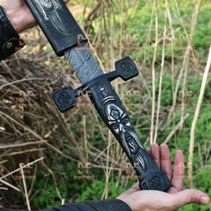 Puede incluir: Una espada negra con intrincadas tallas y una hoja estampada. El mango presenta un diseño detallado de una cara y símbolos de nudos celtas. La espada se sostiene contra un fondo de follaje verde.