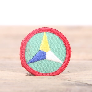 Może przedstawiać: Trójkątna naszywka w kolorze czerwonym, białym i niebieskim z żółtym środkiem. Naszywka jest przyszyta do zielonego tła i ma czerwony obramowanie.