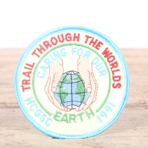 May include: A round, blue and green embroidered patch with the text "Trail Through The Worlds Caring For Our Earth 1991" and a graphic of two hands holding a globe.