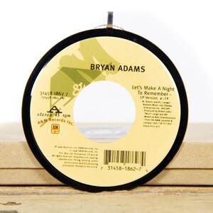 May include: A black vinyl record with a yellow label. The label features the text "BRYAN ADAMS" and the song title "Let's Make A Night To Remember - LP Version 6:19". The record is on a brown surface.