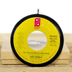 May include: A yellow 45 RPM vinyl record with a black label. The record is titled "Let's Fall in Love All Over Again" by Lou Rawls. The record label includes the song's details, including the record label, the song's length, and the copyright information.