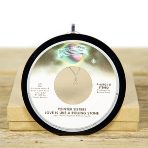 May include: Black and white 45 RPM record ornament featuring a picture of a planet with stars. The record is titled "Love Is Like A Rolling Stone" by the Pointer Sisters. The record label also includes the song's length, the year it was released, and the record label.