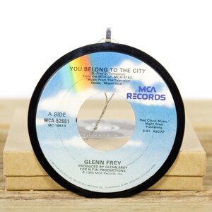 Puede incluir: Un disco de vinilo negro de 45 RPM con una etiqueta blanca. La etiqueta presenta el título de la canción "You Belong to the City" de Glenn Frey, la discográfica "MCA Records" y la información del lado A del disco. El disco está sobre una superficie marrón.
