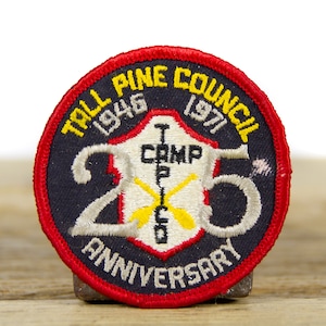 Op de afbeelding: Een geborduurde patch in zwart en wit met rode en gele accenten. De patch heeft de tekst "Tall Pine Council 1946-1971" en "25th Anniversary" rond een schild met de tekst "Camp T.P.C.O."