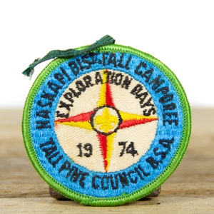 Może przedstawiać: Naszywka haftowana w kolorze niebieskim i zielonym z tekstem "Mackinaw District Fall Campree 1974" i kompasem z żółtym, czerwonym i białym wzorem.