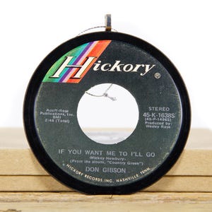 Puede incluir: Un disco de vinilo negro de 45 RPM con una etiqueta colorida que dice "Hickory" y "IF YOU WANT ME TO I'LL GO - Mickey Newbury". El disco es una publicación de Don Gibson y tiene el título de la canción, el nombre del artista y la información del sello discográfico impresa en la etiqueta.