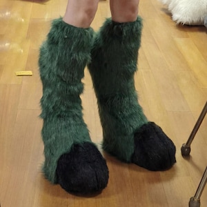 May include: Green furry costume boots with black paw-like feet. The boots are knee-high and made of a shaggy, textured material. The boots are designed to resemble animal legs.