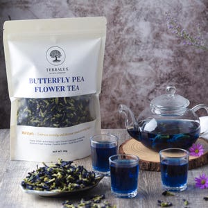 May include: A clear glass teapot and three small glasses filled with blue tea, alongside a packet of Terralux Butterfly Pea Flower Tea. The scene is decorated with purple flowers and a small dish of loose tea.
