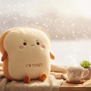 Puede incluir: Un peluche con forma de rebanada de pan tostado con una expresión enfadada, el texto "I'M TOAST" y extremidades pequeñas. Una taza de té floral con vapor, un posavasos y una pequeña planta suculenta están sobre una superficie de madera.