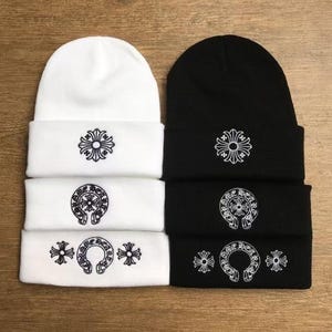 Puede incluir: Dos gorros apilados, uno blanco y otro negro. Cada gorro tiene un borde doblado con un diseño bordado. El gorro blanco tiene tres diseños y el gorro negro tiene tres diseños a juego.