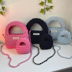 May include: Three sets of crochet handbags in pink, navy blue, and light blue. Each set includes a larger bag and a smaller matching bag, both featuring a top handle and a white label with the text SABY BRAND. Each bag has a chain shoulder strap.