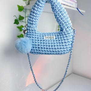 May include: A light blue crochet handbag with a matching pom-pom and silver chain strap. The bag features a label that reads "SABY BRAND". The bag is hanging on a white wall with a shelf and some books.