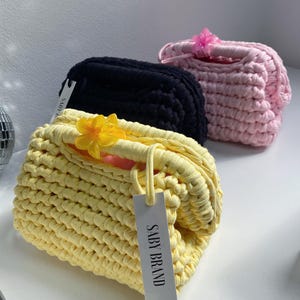 Puede incluir: Tres bolsos de mano de ganchillo hechos a mano en amarillo, negro y rosa. Cada bolso presenta un adorno floral y una etiqueta que dice "SABY BRAND". Los bolsos están hechos de un hilo grueso y texturizado.