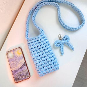 May include: A light blue crocheted crossbody bag with a braided strap, a matching crocheted bow keychain with a silver metal clasp, and a rectangular smartphone case featuring a printed reproduction of a starry night landscape painting.