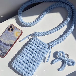 Puede incluir: Una funda de teléfono de macramé azul claro con una correa a juego y un pequeño llavero en forma de lazo. La funda del teléfono es rectangular y tiene una textura tejida. También se ve un teléfono con un diseño de noche estrellada.