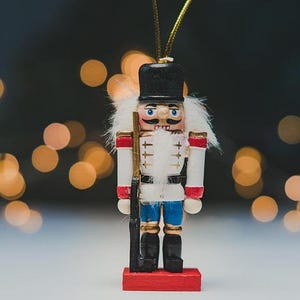 May include: A wooden nutcracker soldier figurine, painted in white, red, black, and blue, stands on a red base. The figurine has a black hat, a white jacket with red accents, and blue pants. It holds a black rifle. The background has blurred golden lights.