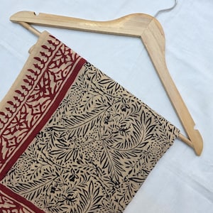 May include: A beige textile with a black and red floral pattern is displayed on a wooden hanger. The textile features a detailed design of leaves and flowers, with a red border. The hanger is made of light-colored wood.