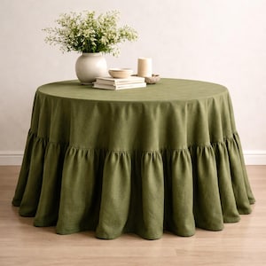 May include: A round olive green tablecloth with a ruffled edge. The table is set with a white vase of flowers, books, a small bowl, and a candle. The tablecloth is made of a textured fabric.